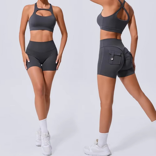 A model is shown from the front and back wearing a dark gray athletic set consisting of a sports bra with a chest cutout and high-waisted shorts with a side cargo pocket, paired with white socks and sneakers on a light gray studio background.