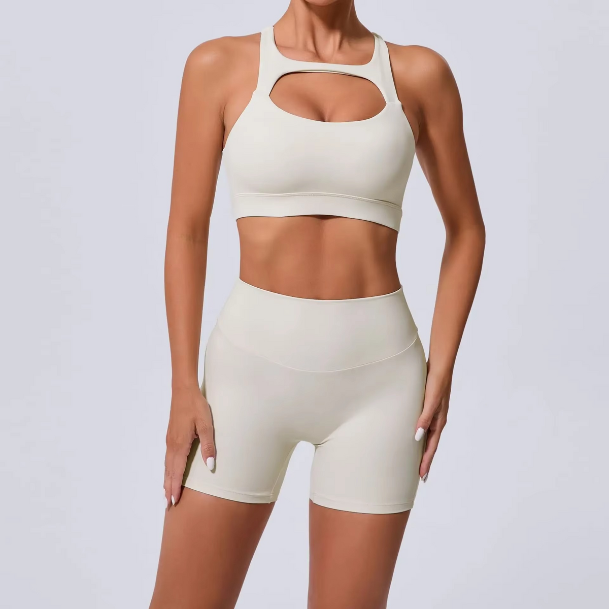 A woman modeling a matching beige activewear set consisting of a sports bra with a chest cutout and high-waisted shorts, against a light gray studio background.