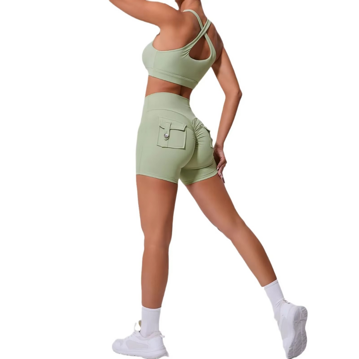 A woman modeling a light sage green athletic set consisting of a sports bra and matching high-waisted cargo shorts with a zippered back pocket, paired with white socks and sneakers against a solid white background.