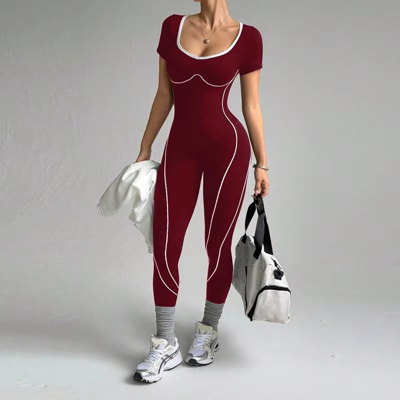 A woman modeling a deep red one-piece athletic jumpsuit with white contrast piping details, styled with gray socks, white sneakers, and carrying a white towel and a white gym bag against a plain gray studio background.